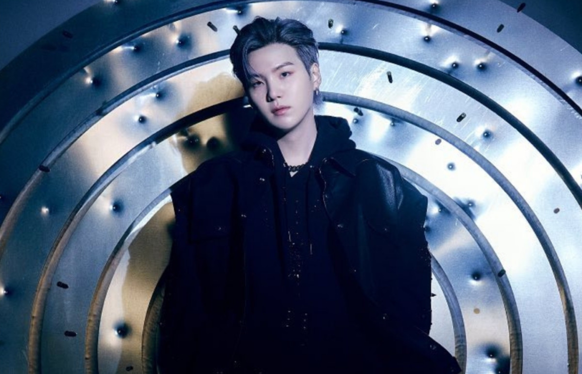 Em fundo metalizado com marcas de bala perdida, SUGA está vestido de preto olhando fixamente para a câmera.