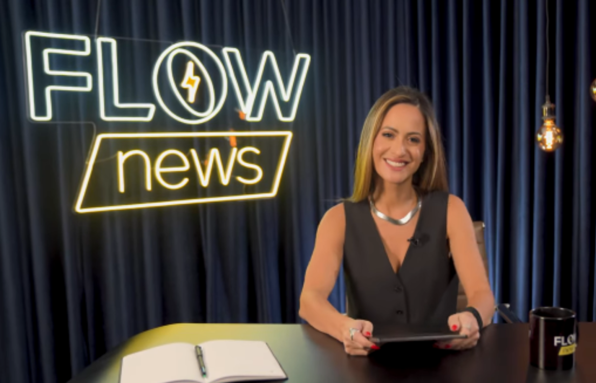 Marcela Rahal sentado em uma mesa com um letreiro luminosos ao lado esquerdo escrito Flow News