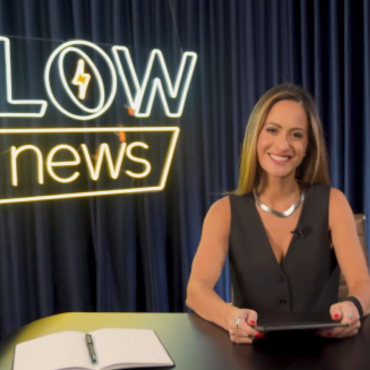 Marcela Rahal sentado em uma mesa com um letreiro luminosos ao lado esquerdo escrito Flow News