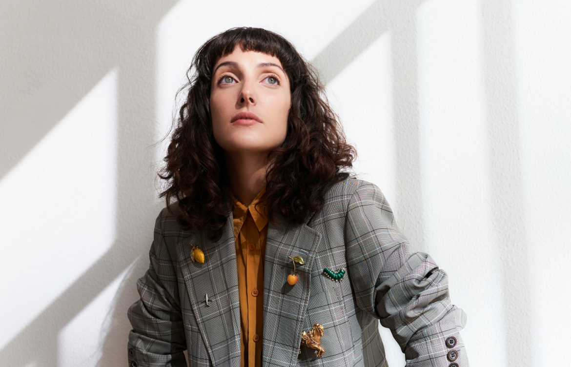 Clarice Falcão vestida com uma jaqueta jeans e uma blusa laranja por baixo diante uma parede branca olhando para um ponto fixo acima da câmera que registra a sua imagem.