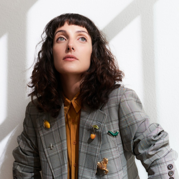 Clarice Falcão vestida com uma jaqueta jeans e uma blusa laranja por baixo diante uma parede branca olhando para um ponto fixo acima da câmera que registra a sua imagem.