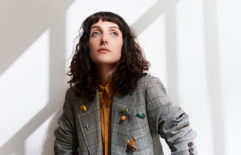 Clarice Falcão vestida com uma jaqueta jeans e uma blusa laranja por baixo diante uma parede branca olhando para um ponto fixo acima da câmera que registra a sua imagem.