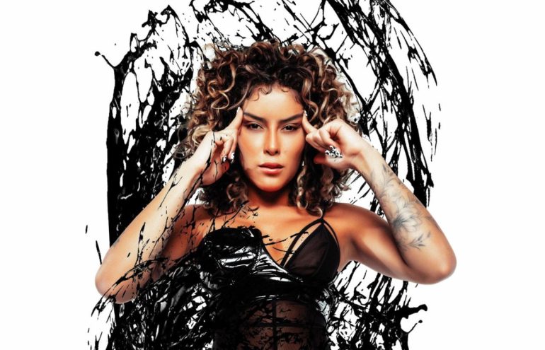 Maia vestida com um vestido preto em uma foto de divulgação esvoaçante enquanto coloca os dois dedos em sua testa.