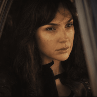 Gal Gadot caracterizada como agente e sentada vestida de preto na janela de um carro.