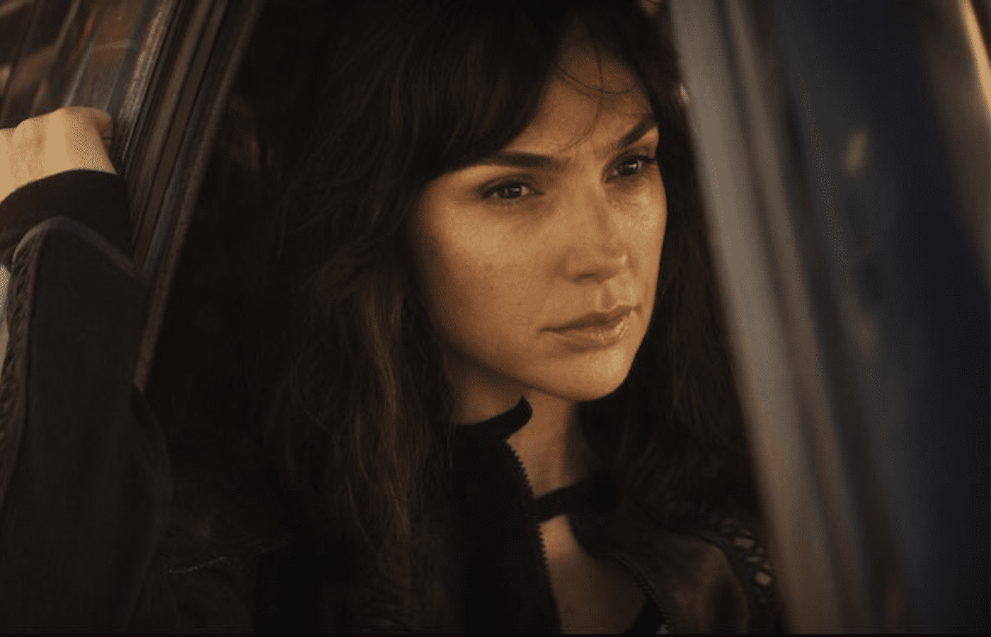 Gal Gadot caracterizada como agente e sentada vestida de preto na janela de um carro.