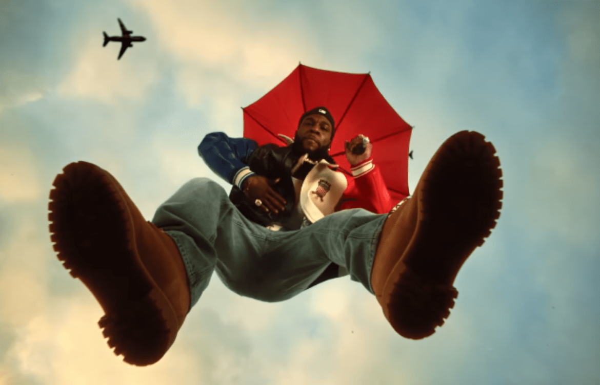 Burna Bou em cima de uma plataforma de vidro. A câmera fima ele de baixo para cima e parece que ele está flutuando. El usa uma bota, calça jeans de cor azul clara e uma jaqueta metade vermelha e outra azul.