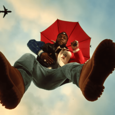 Burna Bou em cima de uma plataforma de vidro. A câmera fima ele de baixo para cima e parece que ele está flutuando. El usa uma bota, calça jeans de cor azul clara e uma jaqueta metade vermelha e outra azul.