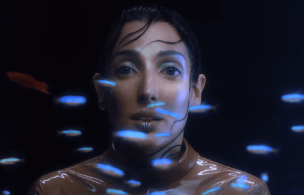 Clarice Falcão canta em um ambiente escuro. na sua frente borboletas na cor azul criadas na pós-produção passam pela sua frente.