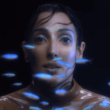 Clarice Falcão canta em um ambiente escuro. na sua frente borboletas na cor azul criadas na pós-produção passam pela sua frente.