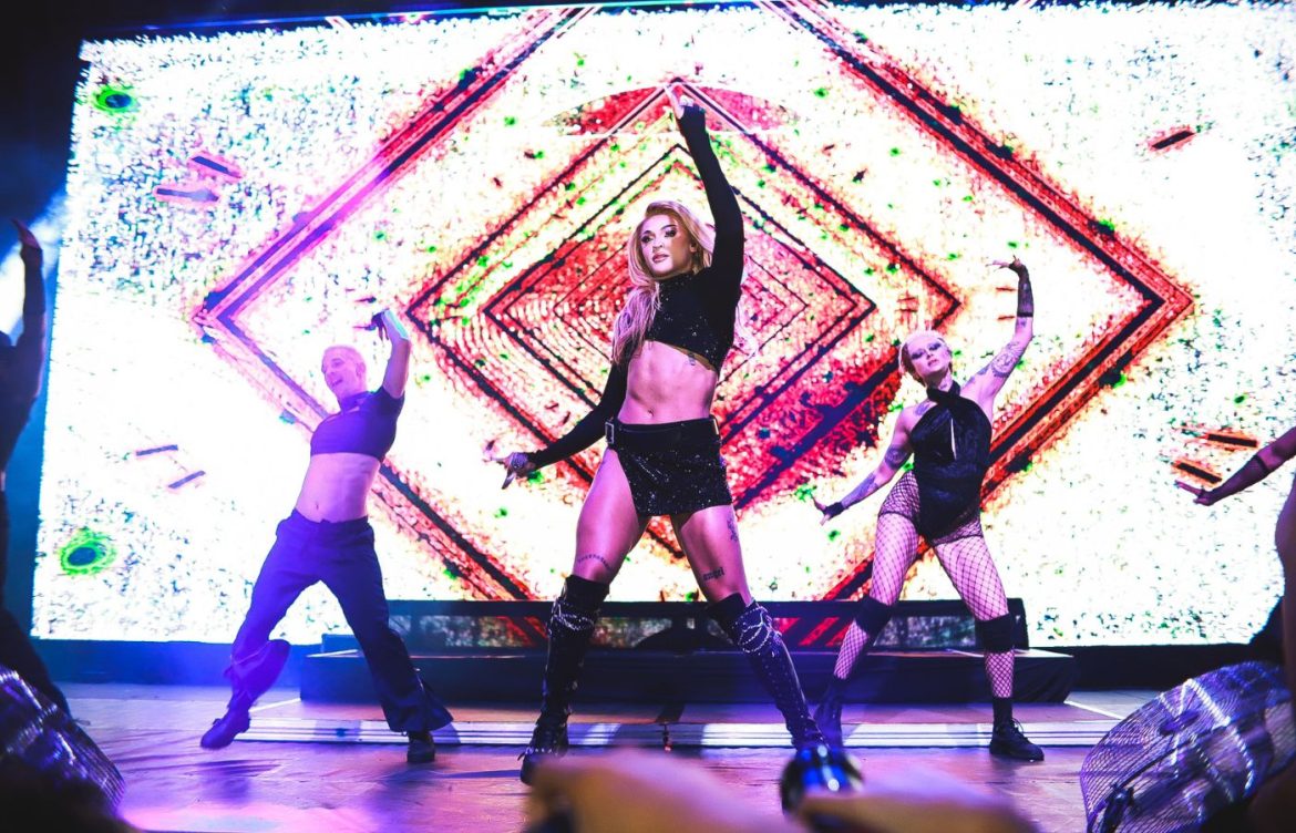 Pabllo Vittar com a mão esquerda para cima enquanto dança uma das coreógrafias de suas canções. Ela está vestida de uma saia preta e top preto, semelhante ao seus dançarinos