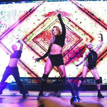 Pabllo Vittar com a mão esquerda para cima enquanto dança uma das coreógrafias de suas canções. Ela está vestida de uma saia preta e top preto, semelhante ao seus dançarinos