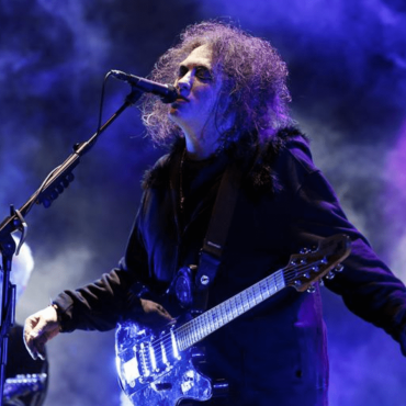Robert Smith, vocalista do "The Cure" de braços aberto vestindo uma blusa de frio de cor preta canta de frente para um pedestal equipado com um microfone. Ele possui uma guitarra presa com uma fita ao pescoço.