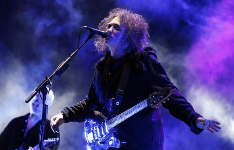 Robert Smith, vocalista do "The Cure" de braços aberto vestindo uma blusa de frio de cor preta canta de frente para um pedestal equipado com um microfone. Ele possui uma guitarra presa com uma fita ao pescoço.