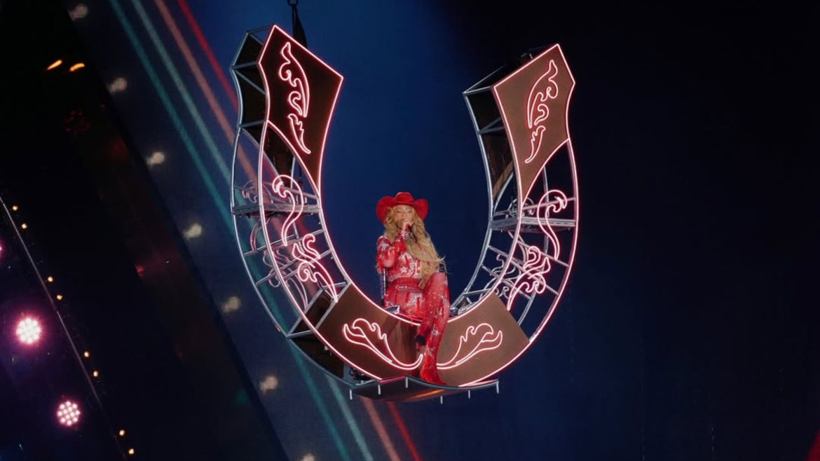 A imagem mostra a cantora Beyoncé durante uma apresentação. Ela está suspensa no ar, sentada em uma estrutura em formato de ferradura gigante, iluminada com luzes de neon rosa. Beyoncé veste um traje brilhante inteiramente vermelho, composto por um body com detalhes reluzentes, botas de salto alto e um chapéu de caubói combinando. Seu cabelo está solto, comprido e ondulado, caindo sobre os ombros. A cena transmite uma estética country moderna e imponente, com Beyoncé no centro do palco, iluminada por holofotes, em um momento teatral e de destaque no show.