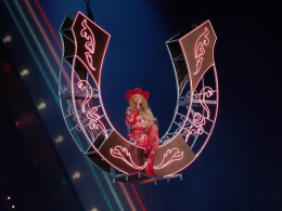A imagem mostra a cantora Beyoncé durante uma apresentação. Ela está suspensa no ar, sentada em uma estrutura em formato de ferradura gigante, iluminada com luzes de neon rosa. Beyoncé veste um traje brilhante inteiramente vermelho, composto por um body com detalhes reluzentes, botas de salto alto e um chapéu de caubói combinando. Seu cabelo está solto, comprido e ondulado, caindo sobre os ombros. A cena transmite uma estética country moderna e imponente, com Beyoncé no centro do palco, iluminada por holofotes, em um momento teatral e de destaque no show.