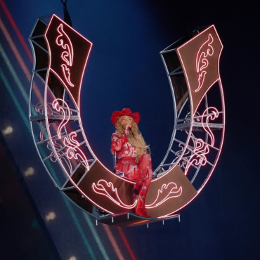 A imagem mostra a cantora Beyoncé durante uma apresentação. Ela está suspensa no ar, sentada em uma estrutura em formato de ferradura gigante, iluminada com luzes de neon rosa. Beyoncé veste um traje brilhante inteiramente vermelho, composto por um body com detalhes reluzentes, botas de salto alto e um chapéu de caubói combinando. Seu cabelo está solto, comprido e ondulado, caindo sobre os ombros. A cena transmite uma estética country moderna e imponente, com Beyoncé no centro do palco, iluminada por holofotes, em um momento teatral e de destaque no show.