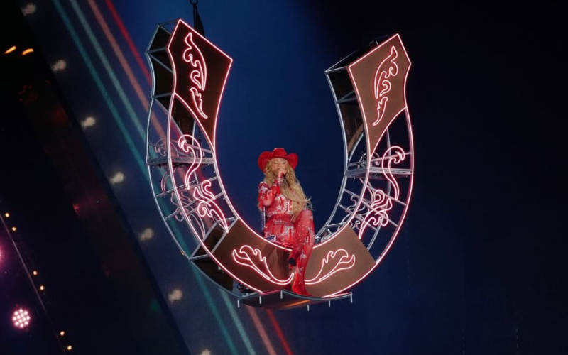 A imagem mostra a cantora Beyoncé durante uma apresentação. Ela está suspensa no ar, sentada em uma estrutura em formato de ferradura gigante, iluminada com luzes de neon rosa. Beyoncé veste um traje brilhante inteiramente vermelho, composto por um body com detalhes reluzentes, botas de salto alto e um chapéu de caubói combinando. Seu cabelo está solto, comprido e ondulado, caindo sobre os ombros. A cena transmite uma estética country moderna e imponente, com Beyoncé no centro do palco, iluminada por holofotes, em um momento teatral e de destaque no show.