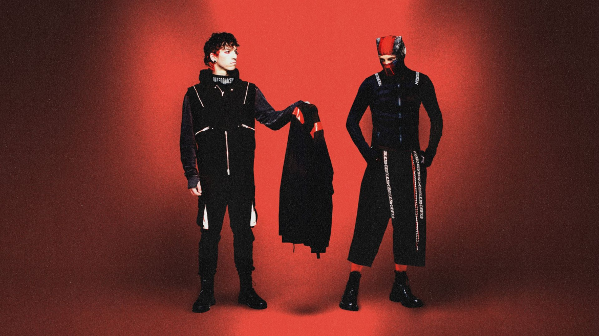 Dois integrantes da banda Twenty One Pilots posam em um fundo vermelho. Um deles, à esquerda, segura uma peça de roupa preta com detalhes vermelhos. Ambos vestem roupas escuras com estilo futurista e elementos gráficos. O integrante à direita usa uma máscara parcial e calças largas, reforçando a estética conceitual da banda.