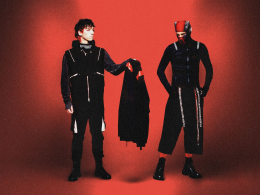 Dois integrantes da banda Twenty One Pilots posam em um fundo vermelho. Um deles, à esquerda, segura uma peça de roupa preta com detalhes vermelhos. Ambos vestem roupas escuras com estilo futurista e elementos gráficos. O integrante à direita usa uma máscara parcial e calças largas, reforçando a estética conceitual da banda.