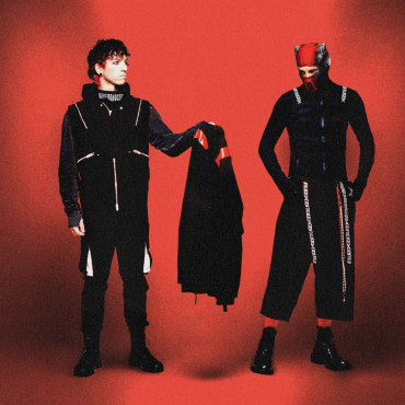 Dois integrantes da banda Twenty One Pilots posam em um fundo vermelho. Um deles, à esquerda, segura uma peça de roupa preta com detalhes vermelhos. Ambos vestem roupas escuras com estilo futurista e elementos gráficos. O integrante à direita usa uma máscara parcial e calças largas, reforçando a estética conceitual da banda.