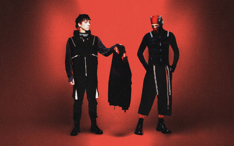 Dois integrantes da banda Twenty One Pilots posam em um fundo vermelho. Um deles, à esquerda, segura uma peça de roupa preta com detalhes vermelhos. Ambos vestem roupas escuras com estilo futurista e elementos gráficos. O integrante à direita usa uma máscara parcial e calças largas, reforçando a estética conceitual da banda.
