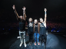 Integrantes da banda My Chemical Romance em pé sobre o palco, de mãos dadas e braços erguidos, agradecendo o público ao final de um show; ao fundo, a plateia lota a arena iluminada com luzes suaves.