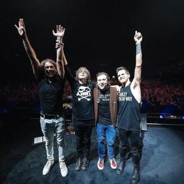 Integrantes da banda My Chemical Romance em pé sobre o palco, de mãos dadas e braços erguidos, agradecendo o público ao final de um show; ao fundo, a plateia lota a arena iluminada com luzes suaves.