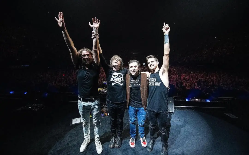 Integrantes da banda My Chemical Romance em pé sobre o palco, de mãos dadas e braços erguidos, agradecendo o público ao final de um show; ao fundo, a plateia lota a arena iluminada com luzes suaves.