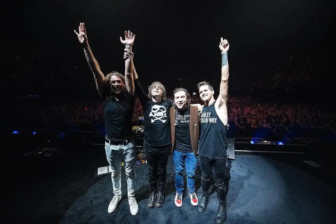 Integrantes da banda My Chemical Romance em pé sobre o palco, de mãos dadas e braços erguidos, agradecendo o público ao final de um show; ao fundo, a plateia lota a arena iluminada com luzes suaves.