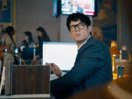 Clark Kent sentado em sua mesa no Planeta Diário, usando terno escuro e óculos, olhando para o lado com expressão séria; ao fundo, colegas Ftoe uma TV exibe um noticiário.