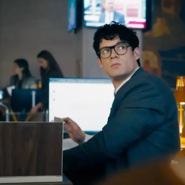 Clark Kent sentado em sua mesa no Planeta Diário, usando terno escuro e óculos, olhando para o lado com expressão séria; ao fundo, colegas Ftoe uma TV exibe um noticiário.