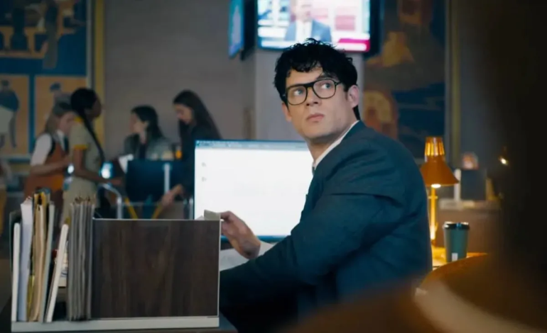 Clark Kent sentado em sua mesa no Planeta Diário, usando terno escuro e óculos, olhando para o lado com expressão séria; ao fundo, colegas Ftoe uma TV exibe um noticiário.