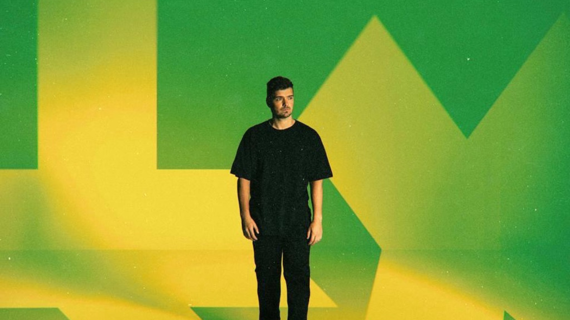 Martin Garrix em pé contra um fundo com as cores verde e amarelo estilizadas, remetendo à bandeira do Brasil. Ele veste camiseta e calça pretas, com expressão séria e olhar voltado para a direita.