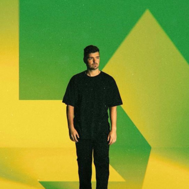 Martin Garrix em pé contra um fundo com as cores verde e amarelo estilizadas, remetendo à bandeira do Brasil. Ele veste camiseta e calça pretas, com expressão séria e olhar voltado para a direita.