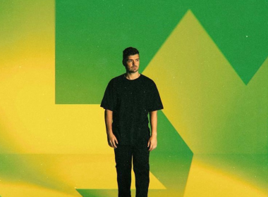 Martin Garrix em pé contra um fundo com as cores verde e amarelo estilizadas, remetendo à bandeira do Brasil. Ele veste camiseta e calça pretas, com expressão séria e olhar voltado para a direita.