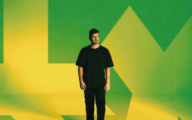 Martin Garrix em pé contra um fundo com as cores verde e amarelo estilizadas, remetendo à bandeira do Brasil. Ele veste camiseta e calça pretas, com expressão séria e olhar voltado para a direita.
