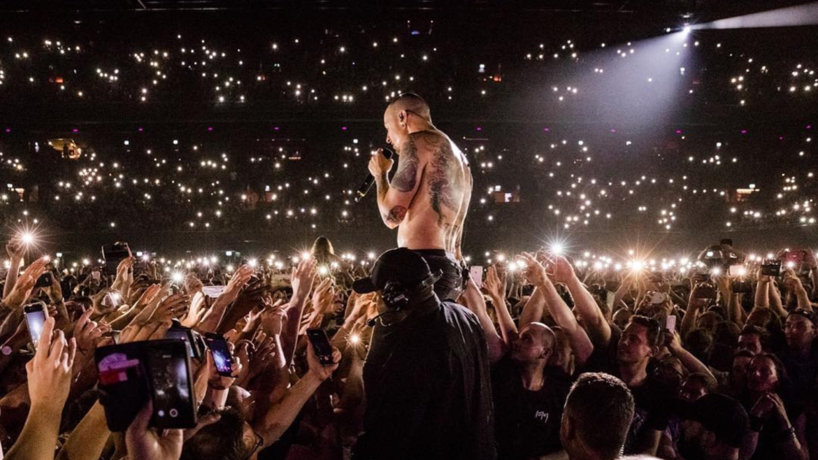 Chester Bennington, vocalista do Linkin Park, canta sem camisa em meio ao público durante show lotado, com milhares de luzes de celulares acesas iluminando a plateia.