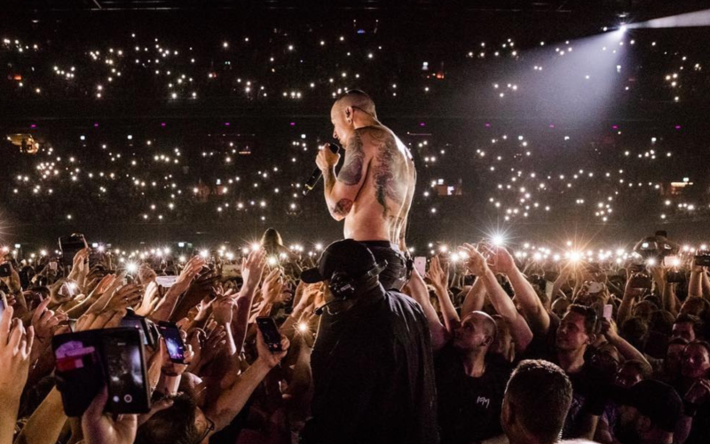 Chester Bennington, vocalista do Linkin Park, canta sem camisa em meio ao público durante show lotado, com milhares de luzes de celulares acesas iluminando a plateia.