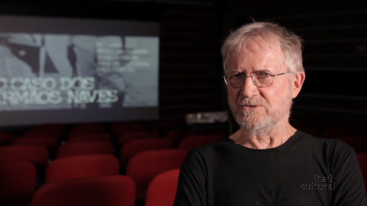 Jean‑Claude Bernardet sentado em uma sala de cinema vazia, usando camiseta preta e óculos, com a tela ao fundo exibindo o título “O Caso dos Irmãos Naves”. Logotipo do Itaú Cultural no canto inferior direito.