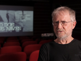 Jean‑Claude Bernardet sentado em uma sala de cinema vazia, usando camiseta preta e óculos, com a tela ao fundo exibindo o título “O Caso dos Irmãos Naves”. Logotipo do Itaú Cultural no canto inferior direito.