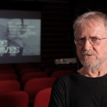Jean‑Claude Bernardet sentado em uma sala de cinema vazia, usando camiseta preta e óculos, com a tela ao fundo exibindo o título “O Caso dos Irmãos Naves”. Logotipo do Itaú Cultural no canto inferior direito.