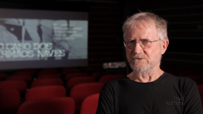 Jean‑Claude Bernardet sentado em uma sala de cinema vazia, usando camiseta preta e óculos, com a tela ao fundo exibindo o título “O Caso dos Irmãos Naves”. Logotipo do Itaú Cultural no canto inferior direito.