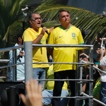 Silas Malafaia discursa ao microfone ao lado de Jair Bolsonaro em cima de um trio elétrico durante manifestação, ambos vestindo camisas amarelas da seleção brasileira, com palmeiras ao fundo e pessoas filmando com celulares.