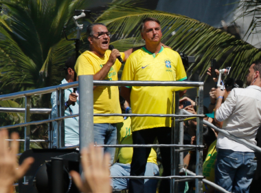 Silas Malafaia discursa ao microfone ao lado de Jair Bolsonaro em cima de um trio elétrico durante manifestação, ambos vestindo camisas amarelas da seleção brasileira, com palmeiras ao fundo e pessoas filmando com celulares.
