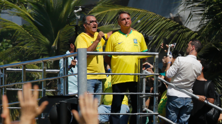 Silas Malafaia discursa ao microfone ao lado de Jair Bolsonaro em cima de um trio elétrico durante manifestação, ambos vestindo camisas amarelas da seleção brasileira, com palmeiras ao fundo e pessoas filmando com celulares.