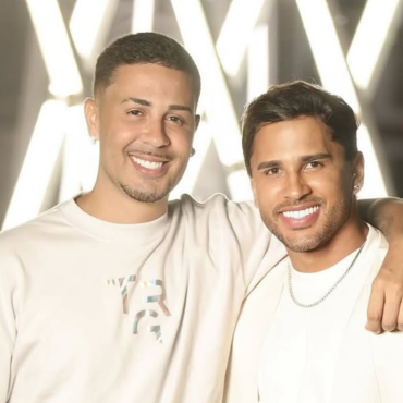 Carlinhos Maia e Lucas Guimarães posam sorridentes lado a lado, com os braços entrelaçados, em frente a um fundo iluminado com luzes brancas em forma de "X". Ambos usam roupas claras e exibem expressão de felicidade.