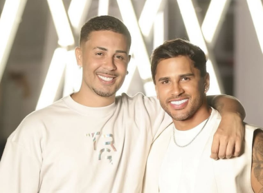 Carlinhos Maia e Lucas Guimarães posam sorridentes lado a lado, com os braços entrelaçados, em frente a um fundo iluminado com luzes brancas em forma de "X". Ambos usam roupas claras e exibem expressão de felicidade.