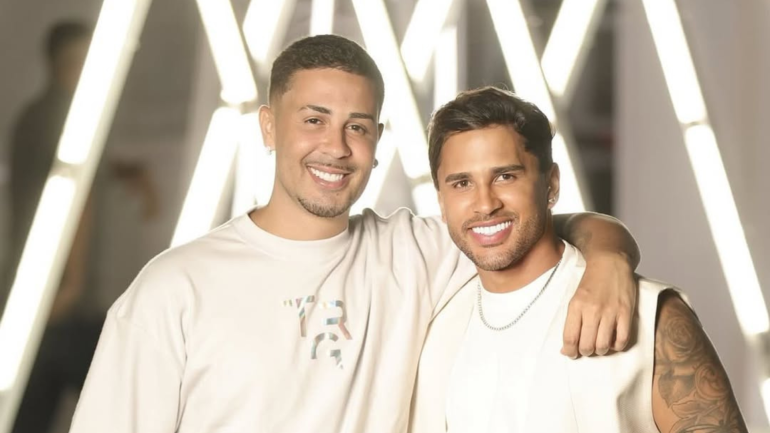 Carlinhos Maia e Lucas Guimarães posam sorridentes lado a lado, com os braços entrelaçados, em frente a um fundo iluminado com luzes brancas em forma de "X". Ambos usam roupas claras e exibem expressão de felicidade.