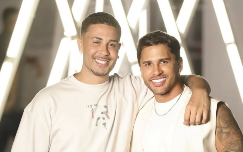 Carlinhos Maia e Lucas Guimarães posam sorridentes lado a lado, com os braços entrelaçados, em frente a um fundo iluminado com luzes brancas em forma de "X". Ambos usam roupas claras e exibem expressão de felicidade.