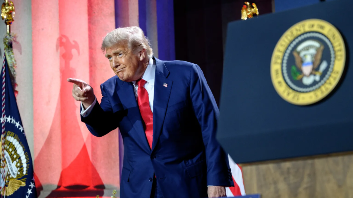 Donald Trump, vestindo terno azul e gravata vermelha, aponta com o dedo indicador durante um discurso. Ao fundo, colunas iluminadas em vermelho e azul e o selo presidencial dos Estados Unidos estão visíveis, reforçando o cenário oficial.